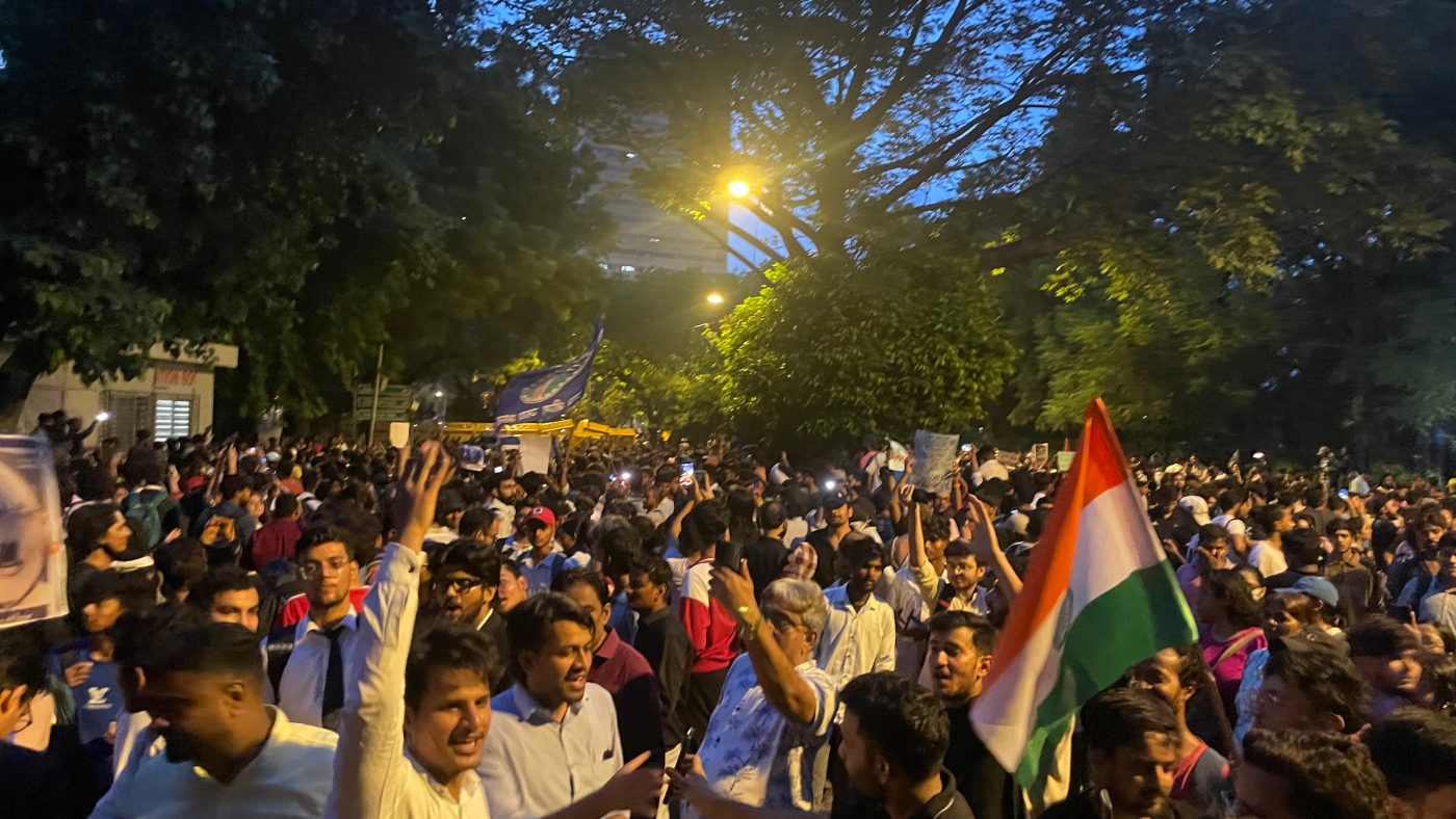A dense crowd of protesters at Jantar Mantar in Delhi at dusk, with placards held up and an Indian flag raised above them.